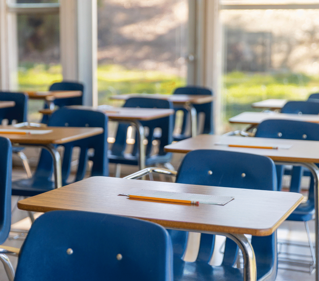 A photo of inside a classroom with no people. There are rows of blue seats with wooden top desks all facing the same direction. There are windows in the back that look onto greenery.