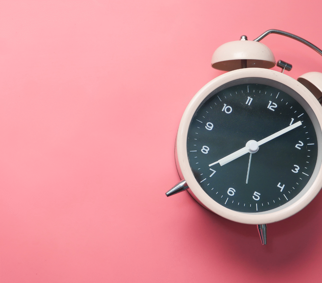 A round shaped, pale pink alarm clock is to the right of the photo and at a tilted angle. The alarm clock is against a pink background.