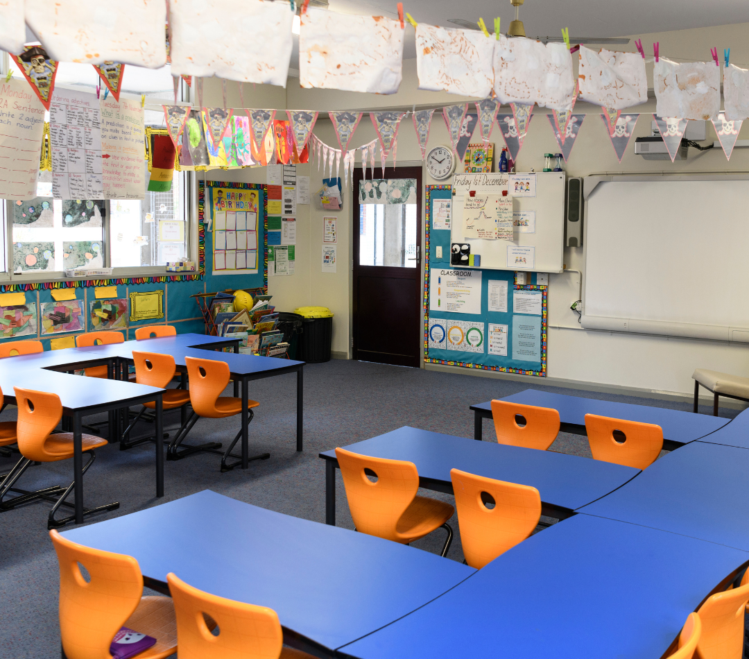 Interior of a primary classroom. There are blue desks, orange chairs and multi-coloured display boards.