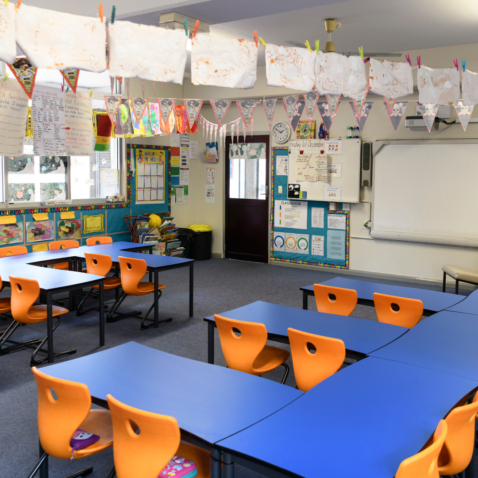Interior of a primary classroom. There are blue desks, orange chairs and multi-coloured display boards.